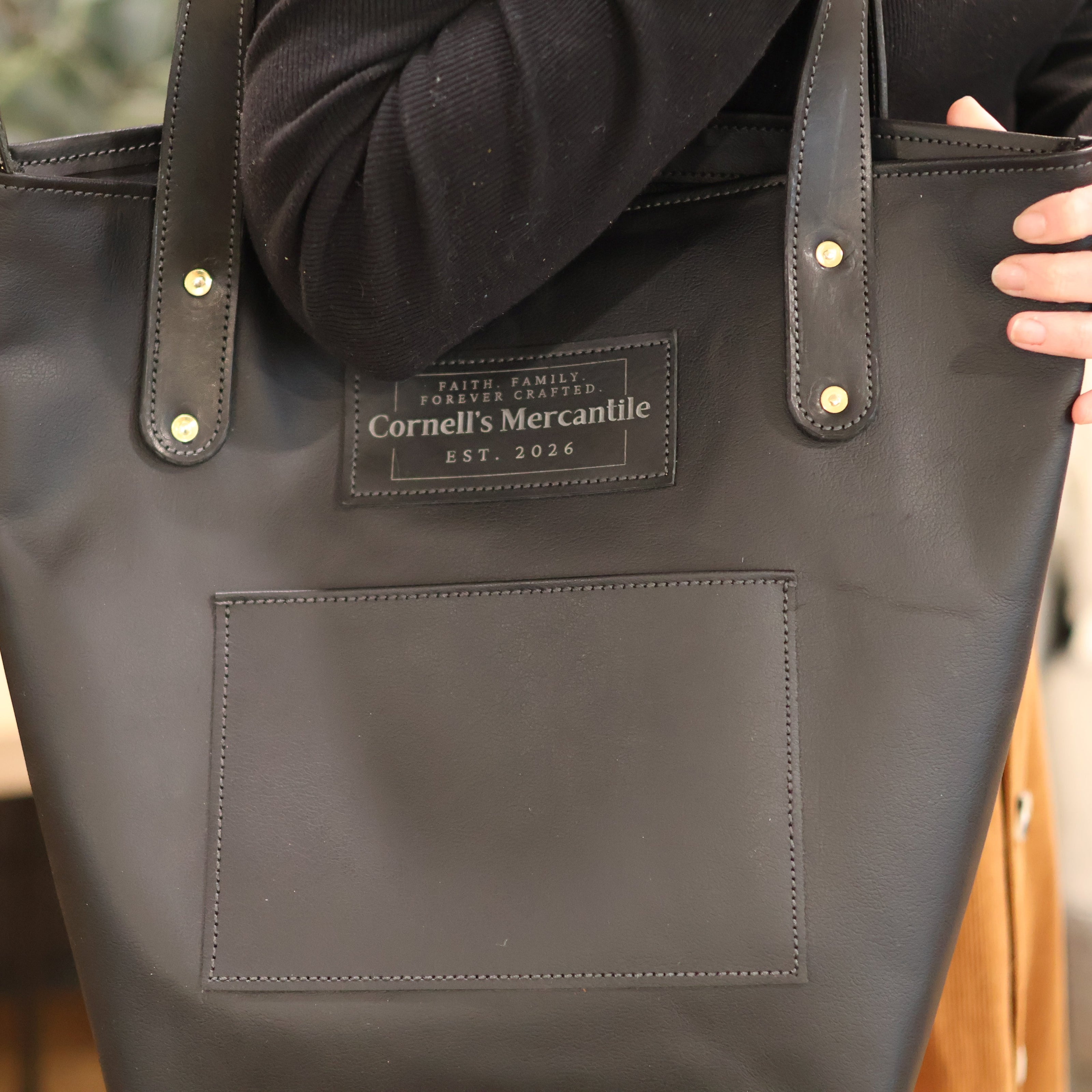 Black tote bag with 'Cornell's Mercantile' label held by a person.